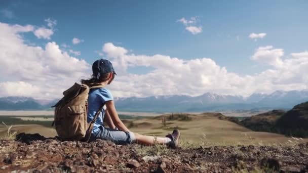 το παιδί σε ένα πεζοπορώ. ένα κοριτσάκι είναι ανάπαυση και θαυμάζοντας τη θέα στα βουνά που κάθεται πάνω στα βράχια. μικρό τουριστικό με ένα σακίδιο - Πλάνα, βίντεο
