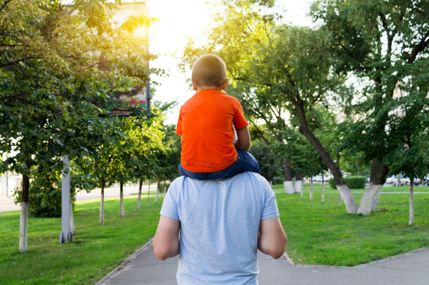 オレンジ色のジャケットの少年は父親の肩の上に座っています。信頼関係の概念. - 写真・画像