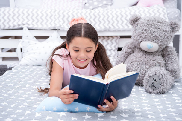Read fairytale in bed. Girl child lay bed with teddy bear read book. Kid prepare to go to bed. Time for evening fairytale. Girl kid long hair cute pajamas relax and read fairytale book to bear toy - Fotó, kép