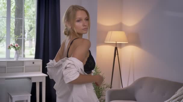 Young sexy blonda lady in lingerie is standing and watching her body in front of mirror, touching bra - Πλάνα, βίντεο