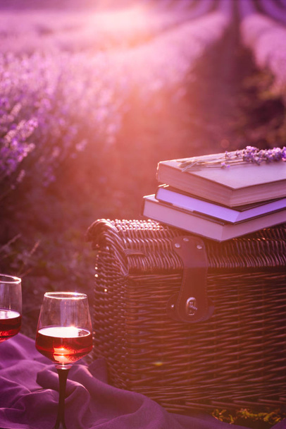 Piknikový koš, staré knihy, sklenice vína v paprscích zapadajícího slunce. Romantický piknik koncepce při západu slunce v voňavé levandule pole. - Fotografie, Obrázek