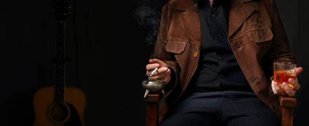 Panorama with the figure of a musician in a suede jacket with a cigarette and a glass of whiskey on the background of acoustic guitar - Photo, image
