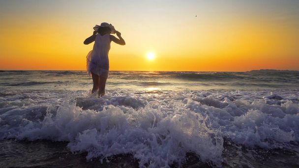 夕日を背景にビーチで楽しんでいる帽子と白いドレスの女 - 写真・画像