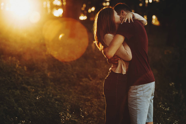 Side view of casual loving couple embracing while standing in green park with glowing sunset light - Foto, Imagem