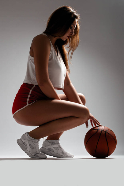 Jovencita sexy en pantalones cortos rojos y top blanco, sentada en estudio con baloncesto. Chica con piernas largas. Mujeres deportistas felices
 - Foto, imagen