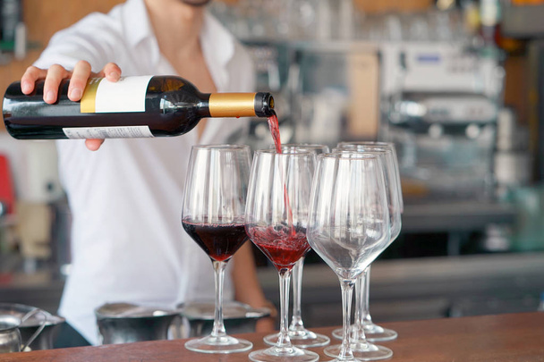 Ένα μπάρμαν άνδρες στέκεται, χύνει κόκκινο κρασί σε ποτήρι από ένα μπουκάλι. Ράφια με τα μπουκάλια αλκοόλ στο παρασκήνιο. Επικεντρώνονται στη γυάλινη επιφάνεια του. - Φωτογραφία, εικόνα