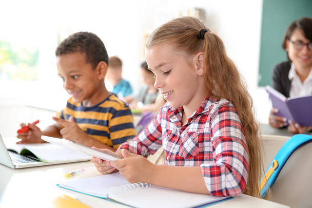 Lindos niños pequeños con aparatos sentados en el escritorio en el aula. Escuela primaria
 - Foto, imagen