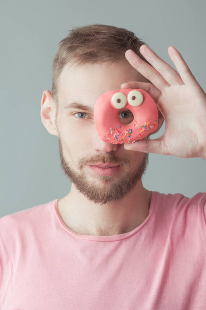 Colour obsession concept. Young handsome hipster guy with beard looking through pink glazed donut over gray background. Minimalistic style. Close up. Studio shot - Valokuva, kuva