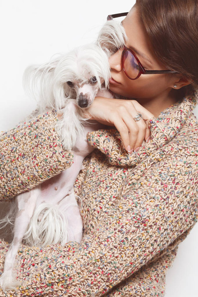 Best girlfriends concept. Young gorgeous lady with natural make-up wearing trendy clothes, eyeglasses kissing small chinese crested dog over white background. Street fashion style. Indoor shot - Fotoğraf, Görsel