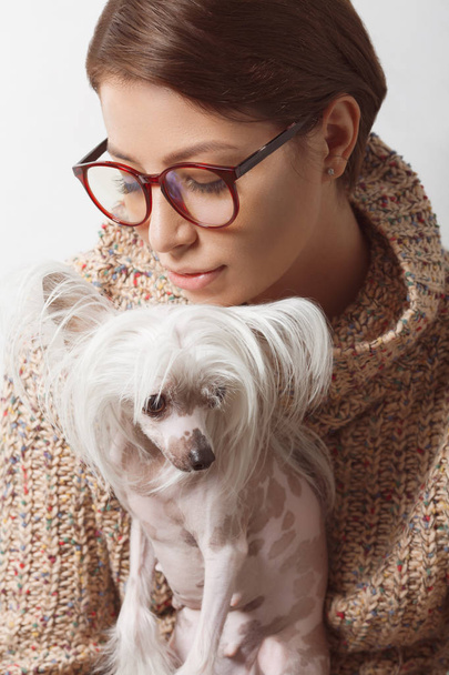 Best girlfriends concept. Young gorgeous lady with natural make-up wearing trendy clothes, eyeglasses kissing small chinese crested dog over white background. Street fashion style. Indoor shot - Foto, Imagen