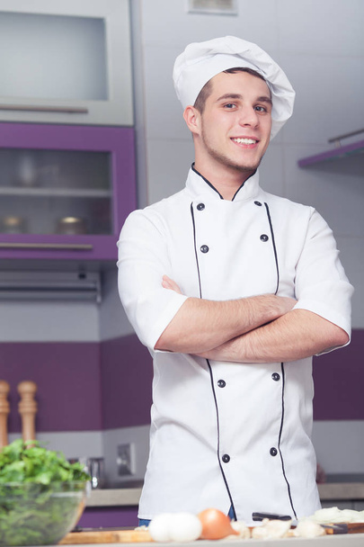 Portrait of funny smiling man in cook uniform  with crossed hands posing in modern kitchen. Indoor shot - Foto, afbeelding