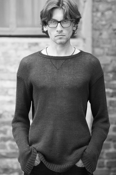 Street fashion concept. Portrait of romantic charismatic man wearing stylish eyeglasses, knitted sweater. Monochrome, black and white outdoor shot - Фото, изображение