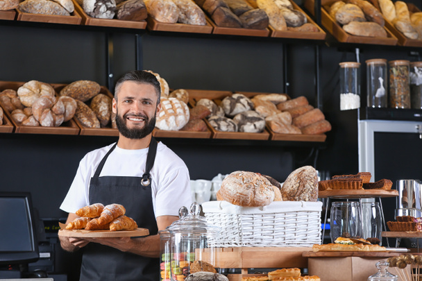 Male baker holding wooden board with delicious croissants in shop - Photo, image