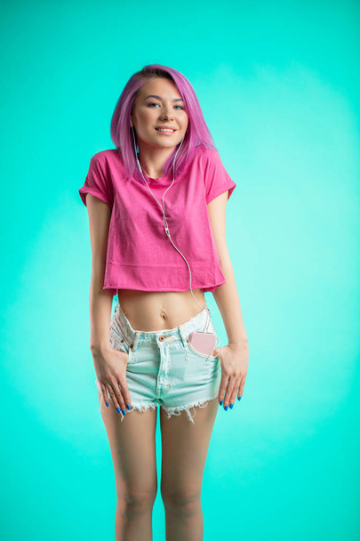 Shy lady with pink hair posing over blue background listening music in headphones. - Foto, immagini