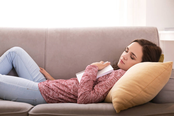 Mujer joven y cansada con libro durmiendo en el sofá en casa
 - Foto, imagen