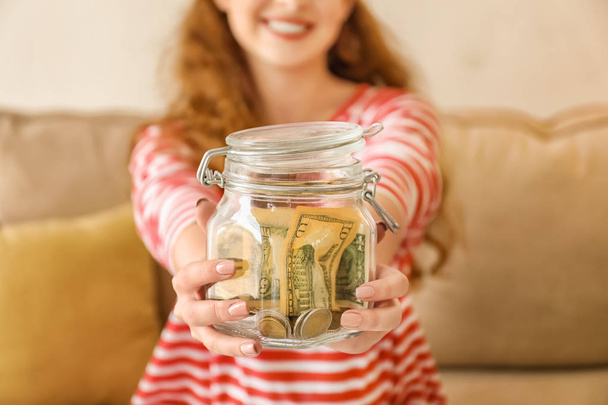 Young woman holding glass jar with dollars indoors. Money savings concept - Photo, Image