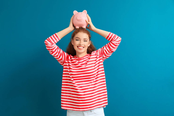 Young woman holding piggy bank on color background. Money savings concept - Photo, Image