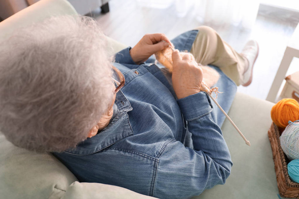 Senior woman sitting on sofa while knitting sweater at home - Foto, immagini