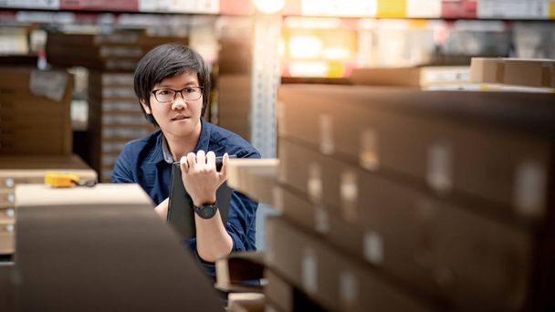 Joven hombre asiático haciendo inventario de producto en caja de cartón en estantes en el almacén mediante el uso de tableta digital. concepto de recuento de inventario físico
 - Foto, imagen