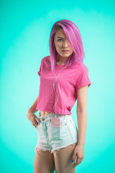 Shy lady with pink hair posing over blue background listening music in headphones. - Φωτογραφία, εικόνα