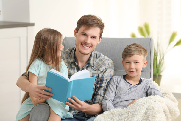 Isä ja hänen lapsensa lukevat kirjaa yhdessä kotona
 - Valokuva, kuva