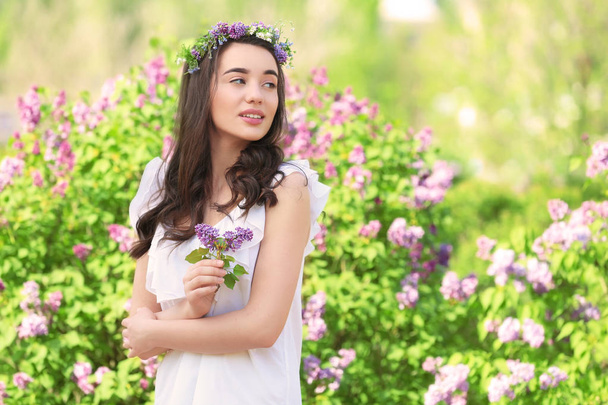 Όμορφη νεαρή γυναίκα που φοράει στεφάνι λουλουδιών κοντά ανθοφορίας θάμνων στην ηλιόλουστη ημέρα άνοιξη - Φωτογραφία, εικόνα