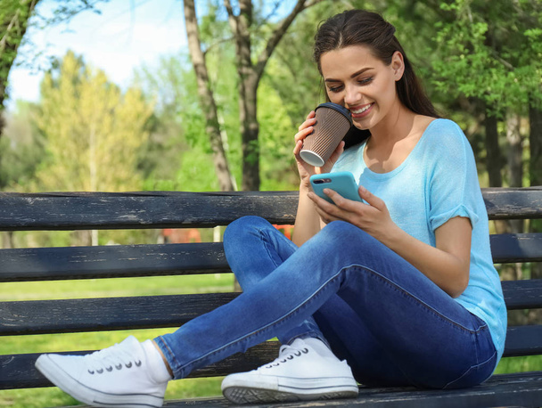Όμορφη νεαρή γυναίκα που αναπαύεται στο πάρκο με φλιτζάνι καφέ και κινητό τηλέφωνο - Φωτογραφία, εικόνα