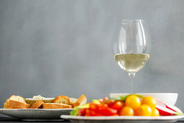 Glass white wine with vegetable snacks and bread croutons against gray concrete wall - Фото, изображение
