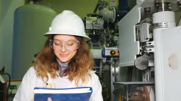 A lány mérnök egy fehér sisak és szemüveg rekordok az adatok egy magazin, úgy néz ki, a kamera és a mosoly, a háttérben a berendezések ipari gáz. - Felvétel, videó