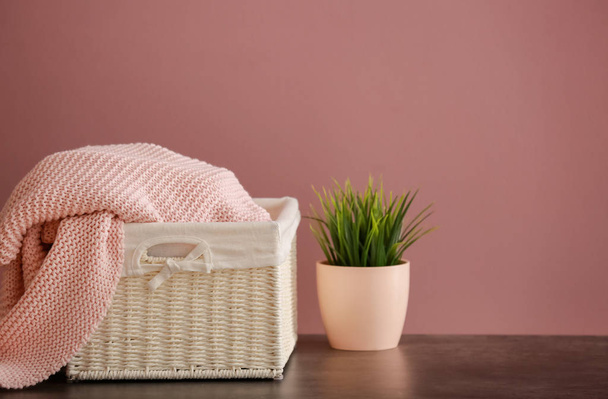 Basket with knitted plaid and plant on table against color background - Фото, зображення