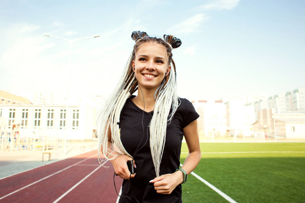 A young woman with African pigtails in her hair runs along the treadmill in a sporty uniform with headphones and listens to music - Fotografie, Obrázek