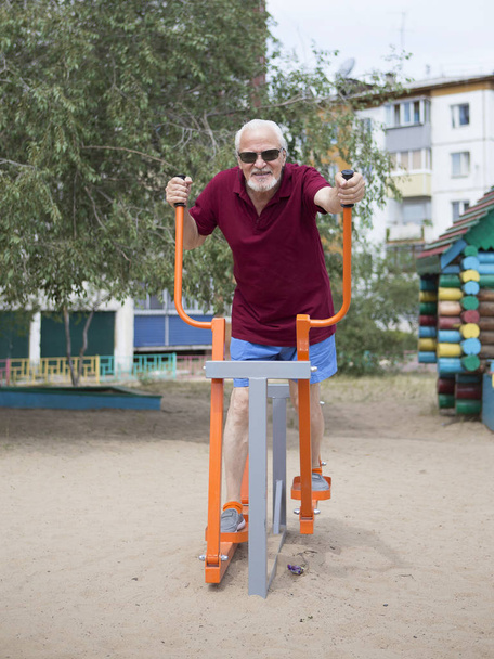Attractive senior man trains on sporting equipment in a city in the open air. The concept of a healthy lifestyle and accessibility of sports training for every person. Available sports equipment in a public place. - Photo, Image