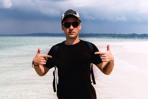 Een toerist met een pet en een zonnebril. Gelukkige man die zich aangeeft tegen de achtergrond van het zeelandschap op een exotisch eiland. kopieerruimte, kopieerruimte - Foto, afbeelding