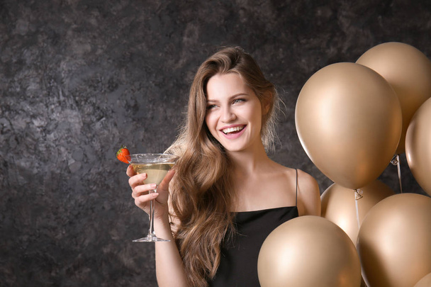 Beautiful young woman with balloons and cocktail on dark grey background - Fotoğraf, Görsel