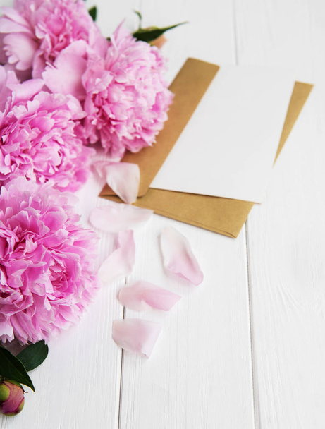 davetiye, zanaat zarf ve pembe şakayık çiçekleri Beyaz ahşap bir masa üzerinde - Fotoğraf, Görsel