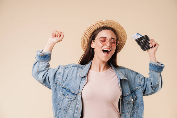 Portrait of a satisfied young brunette girl in straw hat and sunglasses holding passport with flying tickets and celebrating isolated over white background - Photo, Image