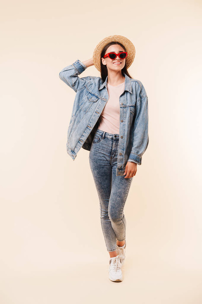 Full length portrait of a smiling young brunette girl in straw hat and denim jacket posing while standing and looking at camera isolated over white background - Foto, immagini