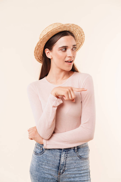 Portrait of a cheerful young brunette girl in straw hat pointing finger away isolated over white background - Фото, зображення