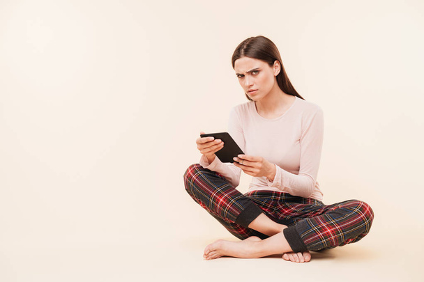 電子ブックを読みながら足で座っているピンクの背景渡った分離のパジャマに身を包んだ深刻な若いブルネットの少女 - 写真・画像