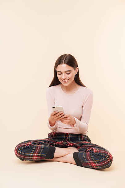 Portrait of a happy young brunette girl using mobile phone while sitting with legs crossed isolated over pink background - Foto, Imagen