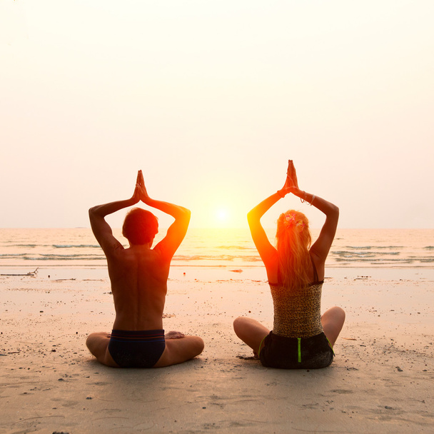 Young couple sitting on the beach of the sea in the lotus position at sunset - Photo, Image