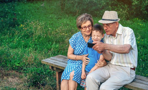 Дедушка делает селфи с бабушкой и внуком сидящими на скамейке
 - Фото, изображение