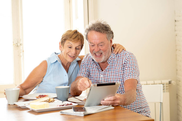 Feliz atractiva pareja de ancianos utilizando tableta de navegación en Internet mientras desayuna y bebe café en casa
. - Foto, Imagen