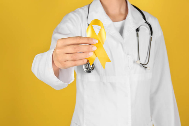 Doctor with yellow ribbon on color background. Cancer awareness concept - Photo, Image