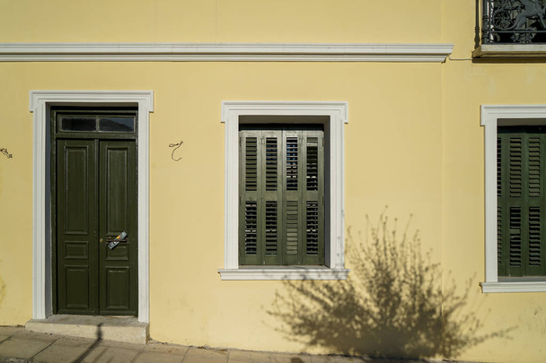 Scene of beautiful urban building facade background in pastel cream yellow plaster paint wall, olive green wooden entry door and windows with tree shadow, Athens, Greece - Photo, Image