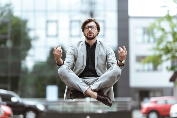attraktiver junger Geschäftsmann schwebt in Lotus-Pose in der Luft und meditiert auf dem Parkplatz - Foto, Bild