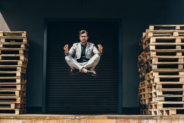 joven hombre de negocios guapo flotando en el aire en pose de loto y meditando delante de la puerta de rodillos
  - Foto, imagen