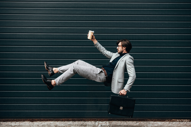 vista lateral de joven hombre de negocios cayendo con taza de papel de café delante de la puerta de rodillos
  - Foto, imagen