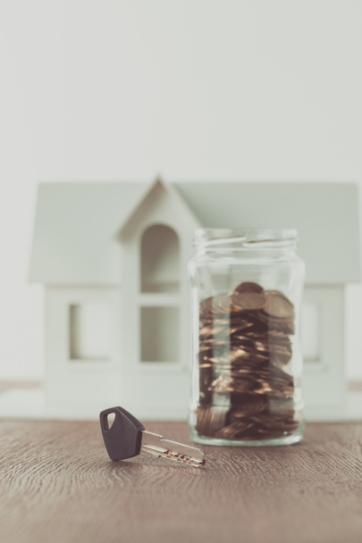 key near jar of coins on tabletop with small house on background, saving concept - Photo, Image