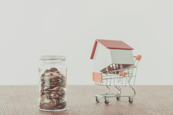small wooden house on supermarket cart, jar of coins on wooden table, saving concept - Photo, Image